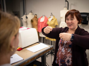 A female teacher instructing a blonde student in a costume shop with mannequins in the background