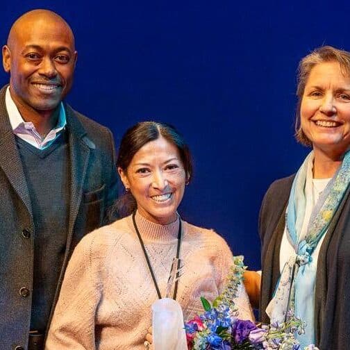 Three people standing on stage, one holding an award and flowers