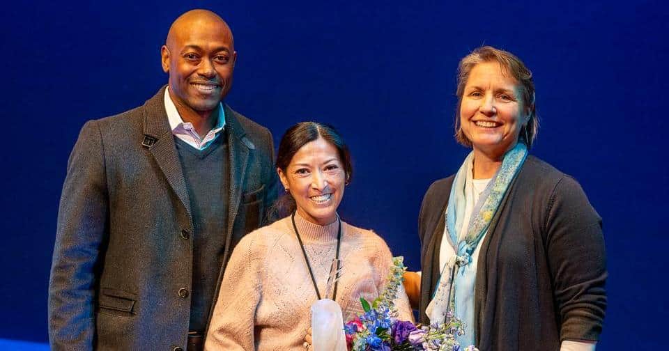 Three people standing on stage, one holding an award and flowers